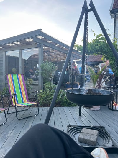 The image depicts an outdoor gathering scene at a private residence in Sandefjord, Norway, likely during a summer evening. A group of people, some wearing motorcycle club attire, are socializing around a fire pit. The environment includes a wooden deck, a greenhouse structure with glass walls, lush green vegetation, and outdoor furniture.
Specifically, a large black fire pit hangs from a tripod structure, with smoke emanating from burning wood within. In the foreground, a person's legs are visible, with a colorful striped deck chair to their left. Behind the fire pit, several individuals are engaged in conversation. One woman, wearing a black t-shirt with "HARLEY-DAVIDSON" printed on it, is looking to the right. Another man in the background is wearing a black vest over a t-shirt.
The greenhouse, to the left of the frame, has wooden framing and transparent roofing, with people visible inside. The overall atmosphere suggests a relaxed and informal social event. The lighting indicates it is either late afternoon or early evening, with the sky overcast.