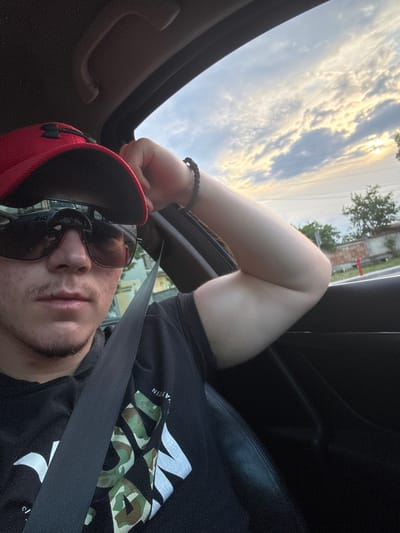 This image, rotated ninety degrees counter-clockwise, features a person seated inside a car, looking towards the camera from an upward angle. The subject, a male with a short goatee, is wearing a red baseball cap and dark sunglasses. A black beaded bracelet adorns his left wrist. He is dressed in a black t-shirt featuring a camouflage and white graphic design, with a black seatbelt diagonally across his chest.
The scene is set inside a vehicle, with a clear view of the sky through what appears to be a sunroof or open window. The sky is filled with scattered clouds, illuminated by golden sunlight, suggesting a late afternoon or early evening time. The sun itself is partially visible, peeking through the clouds in the upper portion of the frame. In the distant background, visible outside the car, are trees and faint outlines of buildings or structures, hinting at a semi-urban environment in Lesnovo, Bulgaria. Subtle power lines are also discernible against the sky. The car's dark interior, including portions of the door frame and ceiling, frames the shot.
On the subject's t-shirt, partially obscured by the seatbelt, are white and camouflage-patterned letters. The sequence "WIN" is clearly visible, with other partial letters suggesting a longer word or phrase that is not fully decipherable. The person's expression is neutral, largely hidden by sunglasses.