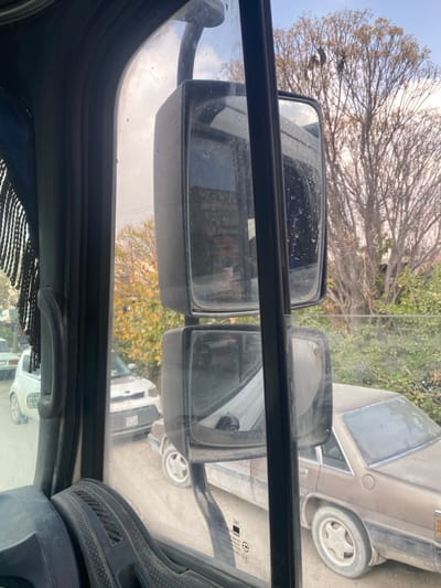 The image presents an exterior view observed from the passenger-side window of a stationary vehicle, identified as a truck, located in Dahiyat Harasta, Syria. The foreground displays a dark grey interior door frame with a black fringed curtain on the left, and a textured portion of the dashboard at the bottom left. Centrally positioned are two stacked, rectangular exterior rearview mirrors, both appearing dusty and smudged. The upper mirror reflects a distant building and indistinct Arabic text, while the lower mirror shows a reflection of a white vehicle. The window glass itself exhibits dirt and water spots.
Outside the vehicle, an unpaved, dusty road or path is visible. Multiple vehicles are parked along this path. To the far left, a white small car, possibly a compact model, is partially visible facing away. Closer to the center, a white vehicle resembling a Kia Soul, with a damaged front bumper and a visible license plate including the numbers "3169," faces towards the viewer. To the right, an older model brown sedan, heavily coated in dust, is parked facing away. The tires and hubcaps of the sedan are also dust-covered. The surrounding environment consists of sparse vegetation, predominantly deciduous trees with mostly bare branches, indicating a cooler season. One tree on the left has visible yellowish-green leaves. The background features a light blue sky with scattered clouds. No individuals are visible, and no dynamic actions are depicted.