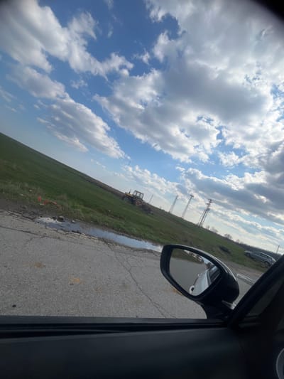 The image depicts an outdoor rural or semi-rural scene in Sredna mahala, Bulgaria, captured from the interior of a vehicle. The perspective is through the passenger-side window, angled upward and to the left.
The sky occupies the upper two-thirds of the frame, displaying a vibrant blue with numerous white and light grey cumulus clouds scattered across it.
In the mid-ground, a wide, undulating green field stretches across the landscape. Positioned within this field, a yellow and orange tractor with an attached implement, possibly for tilling or plowing, is visible. Extending horizontally through the field is a long, dark, elevated ridge or mound of earth. Near the foreground edge of the field, adjacent to the road, an orange piece of discarded material or construction debris is present.
The immediate foreground features a section of an asphalt road with visible cracks, divots, and several irregular puddles reflecting the sky. To the far right, part of a vehicle's black side mirror and the upper portion of a black car door panel are visible. The mirror reflects a portion of the vehicle's white body and a glimpse of the road behind it.
In the background, three distinct tall, lattice-structured metal towers, characteristic of electricity transmission, ascend from the field. Further to the right in the distant background, a grey SUV-style vehicle is parked or stopped on the side of the field or an unpaved track. The horizon also shows a distant line of trees. No individuals are visible in the scene.