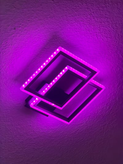 A close-up, angled shot shows a modern, geometric light fixture mounted on a textured, purple-lit wall. The fixture consists of two nested, square-shaped frames, each outlined with a strip of small, bright LED lights that emit a vibrant purple glow. The frames are slightly offset, creating a layered, three-dimensional effect. The wall behind the fixture is illuminated with the same purple light, highlighting its rough, uneven texture. The overall impression is one of contemporary design and atmospheric lighting. This image was taken in Dorsten, Germany.
