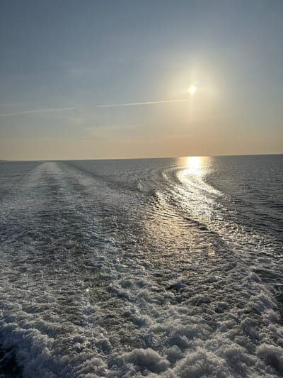 The image presents a stern view from a moving vessel traversing open waters near Palaiopoli, Greece. A prominent, turbulent white wake extends from the foreground towards the horizon, indicating the vessel's propulsion. The sun is positioned in the upper right quadrant of the sky, casting a bright, reflective path of light that shimmers across the water's surface from the middle ground to the horizon line. The sky exhibits a gradient of light blue to pale yellow near the horizon, with two faint, linear contrails visible in the upper left. A low, indistinct landmass is visible along the left horizon. No individuals or specific objects are identifiable on the water or distant land. The overall environment is marine, under a clear sky during daytime.