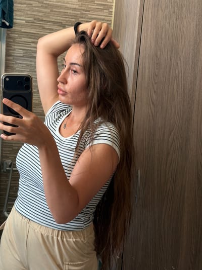 A young woman with long brown hair is taking a selfie in a bathroom. She is wearing a black and white striped t-shirt and light-colored pants. Her left arm is raised, and she is holding her hair. The background shows a tiled wall, a wooden cabinet, and a mirror. The image appears to be taken in a home bathroom in Dromolaxia - Meneou Municipality, Cyprus.