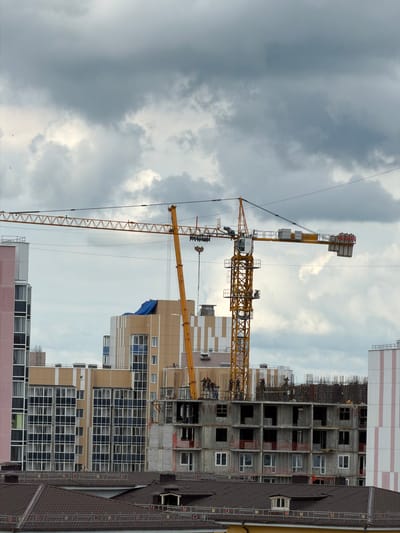 A construction site in Tsiolkovsky, Russia, features a large yellow tower crane with its boom extending horizontally. The crane is positioned in front of several multi-story buildings under construction. One building has a partially covered roof with a blue tarp. Another building under construction shows exposed rebar and concrete structures, with several workers visible on the upper levels. To the left of the crane, a pink and white building with many windows is completed. The sky is overcast with dark, heavy clouds. Below the construction site, several residential buildings with dark brown tiled roofs are visible.