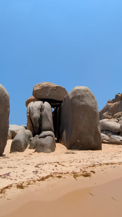 The image depicts a rocky beach in Phuoc Dinh, Vietnam, with large granite boulders clustered together against a clear blue sky. The beach sand is light brown and scattered with seaweed and some litter. The rocks are various shades of grey and tan.