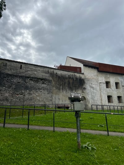 An exterior, ground-level photograph depicts a section of a historical city wall and an attached building under an overcast sky in Schongau, Germany.

The foreground consists of a sloping green lawn, dotted with lighter green clover, leading up to a dark metal railing that traverses horizontally across the frame. Behind the railing, a narrow dark-grey path is visible. A grey metal pole, approximately 1.5 meters tall, stands centrally in the foreground, supporting a rectangular grey electrical junction box. Mounted atop this box is a multi-lamellaed, industrial-style floodlight fixture, facing the structures.

To the left, a tall, weathered dark-grey stone city wall dominates, exhibiting irregular texture, visible mortar, and darker vertical staining. It features small, rectangular openings and a larger, recessed rectangular area in its upper section, topped with a row of small, dark roof tiles.

Attached to the right side of the wall is a multi-story building constructed of light-colored stone or stucco, revealing some exposed brickwork. The building has a steep, light-colored gabled roof to its right and a flatter, rust-red metal roof section extending leftward along the top of the adjacent wall. Multiple rectangular windows with dark frames and stone sills are visible on its facade. A dark wooden bench is positioned against the wall, and to its right, an arched entrance or passageway is discernible below a wall-mounted lantern. No people, active subjects, or interactions are visible.