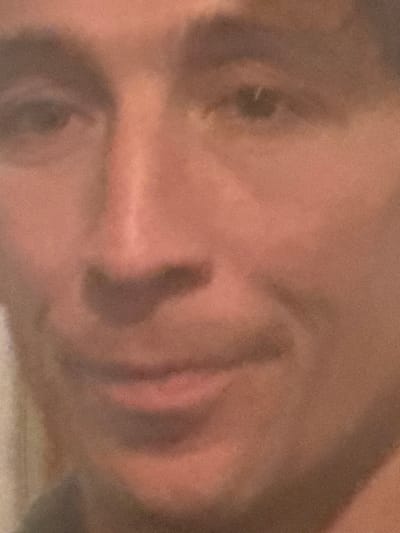 The image displays a close-up, slightly out-of-focus portrait of an adult individual's face, observed in the city of Roanne, France. The subject has light to medium skin tone, dark eyes, and dark, visible eyebrows. A prominent nose is centered in the frame. The mouth shows a subtle, closed-lip smile, and the gaze is directed forward. The background is uniformly indistinct and blurry, lacking any discernible objects, structures, or environmental identifiers. No specific actions or interactions beyond the direct facial presentation are evident.