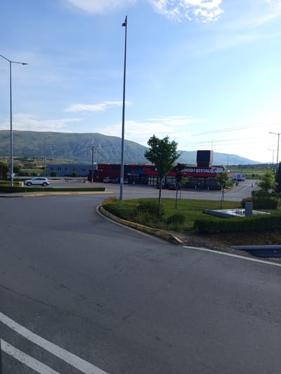 In Dragoman, Bulgaria, on a clear day, a white SUV drives past a row of commercial buildings. One building, marked "TURKISH RESTAURANT," features large windows and outdoor seating. A prominent tall lamppost stands to the right of the scene, and other lampposts dot the landscape. Mountains loom in the background, with a clear blue sky above. The foreground shows a paved road and a well-maintained green area with bushes and trees.
