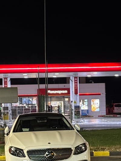 A white Mercedes-Benz is parked at a gas station in Chuchuligovo, Bulgaria, at night. The gas station has a convenience store called "Минимаркет" ("Minimarket"). The gas pumps are illuminated with red lights. The sky is dark.
