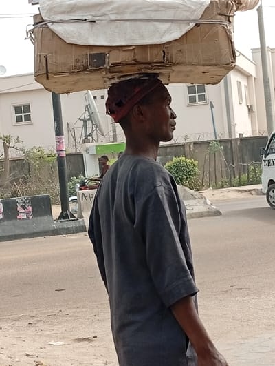 The image displays an outdoor street scene in Darmanawa, Nigeria, during daylight. A dark-skinned individual, likely male, is positioned in the foreground, shown in right profile, carrying a large, rectangular object on their head. The object appears to be constructed from cardboard, topped with a white fabric or tarpaulin, and secured with dark straps; it exhibits wear. Beneath the load, the individual wears a reddish headwrap or padding. They are dressed in a dark grey, short-sleeved tunic-style garment, with their left arm visible beside their body.
In the mid-ground, a paved road stretches horizontally. To the left of the road, a dark grey concrete barrier is visible, affixed with multiple posters featuring pink and white elements. A black utility pole stands directly behind this barrier. Further right on the road, the front section of a white vehicle, possibly a van, displaying partial lettering "Y.H.U." is present.
The background features several light-colored, multi-story buildings with rectangular windows, one of which has an exterior air conditioning unit. A satellite dish is visible between these structures. Sparse green vegetation lines a fence in the background. A blurred figure stands near a green machine, possibly an ATM, in the distance. The immediate foreground shows an unpaved ground area composed of dirt and scattered minor debris.