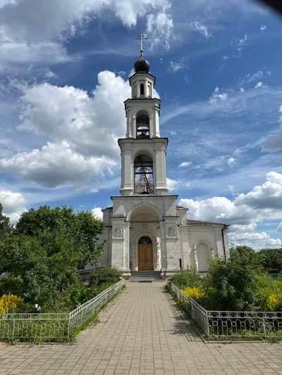 The image captures a bright, sunny day at a religious complex in Ерино, Russia. The primary subjects are a church bell tower and a section of the church building, both constructed from light-colored stone. The bell tower is tall and slender, topped with a dark, domed roof and a cross. It features multiple arched openings where bells would typically hang, and an ornate cross at its apex. Adjacent to the bell tower is the main church structure, also built of stone, with arched windows and decorative architectural details. A wooden door is visible on the church facade, suggesting an entrance.
The scene is set outdoors, with a clear blue sky dominating the upper portion of the image, dotted with fluffy white clouds. Lush green trees and vibrant yellow flowers flank the path leading towards the church. A decorative white metal fence runs along the edge of the path, separating it from the garden area. The ground is paved with light-colored bricks, forming a walkway.
The overall mood is peaceful and serene, characteristic of a quiet day at a place of worship. The bright sunlight and clear sky indicate it is likely midday or early afternoon. There is no visible text in the image.