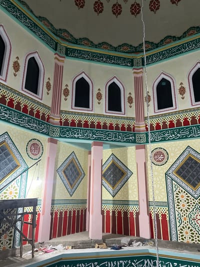 The interior of a mosque features intricate decorative elements. One side exhibits a series of arched windows punctuated by slender columns, all framed by detailed borders with Arabic script and geometric patterns. The opposite side presents diamond-shaped window-like designs. Pillars divide the space, painted with a pale pink hue. The ceiling is adorned with elaborate geometric and floral designs in light colors.