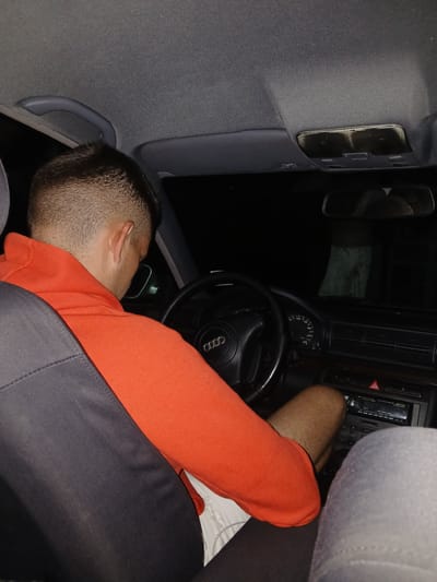 A man is slumped over the steering wheel of a car. The car appears to be an Audi, based on the logo on the steering wheel. The man is wearing an orange shirt. The interior of the car is gray. The image is taken inside the vehicle, facing toward the dashboard and the driver's seat. The car's radio is visible. Location: not found.