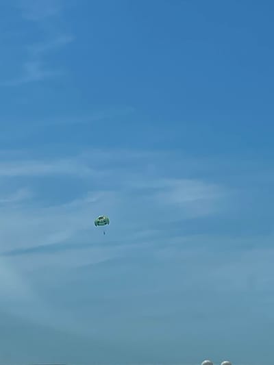 In the city of Lermontovo, Russia, under a bright blue sky with wispy clouds, a person is parasailing high above. The bright green and yellow parachute is clearly visible against the expansive sky. The parasailer appears to be suspended by a thin line. The foreground is obscured by what appears to be the blurred tops of buoys, suggesting the activity is taking place near the water.
