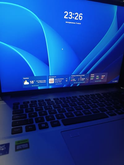 The image shows a close-up view of a laptop screen displaying a Windows 11 desktop. The background is a vibrant blue abstract pattern. On the screen, the time "23:26" is prominently displayed, along with the date "воскресенье, 5 июля" (Sunday, July 5th). To the left, a weather widget shows "Kaushany" with a temperature of "18°C" and a forecast of "Low temps to drop next Wednesday". To the right of the weather widget, an "Event near you" widget displays "Ready Fest Chisinau" with the location "Botanical grad" and the date and time "Saturday, 7:00 PM". Further to the right, a "Trending in World Cup coverage" widget shows results for Brazil, Paraguay, and Norway. Another widget, "Markets", displays information for "WIDOW", "AWGT", and "USD/INR". A portion of the laptop keyboard is visible in the foreground, including the "Caps Lock" and "Ctrl" keys. A sticker for "GeForce RTX 3060" is visible on the lower left of the laptop. The overall lighting suggests a dimly lit environment, casting a blueish hue over the scene. There is no indication of a specific location in Medveja, Moldova.