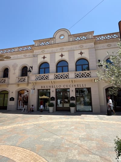 The image shows the exterior of a Brunello Cucinelli store, likely located in La Roca Village, a luxury outlet shopping center in La Roca del Vallès, Spain. The building is a light beige color with architectural details such as arched windows, decorative railings, and ornate friezes. The storefront features large glass windows displaying merchandise and mannequins. The foreground shows a tiled plaza with a circular pattern. Several individuals are visible: two men are standing near a store entrance, another person is sitting on the ground, and a person is walking away with a suitcase. The sky is clear and blue, indicating a sunny day.