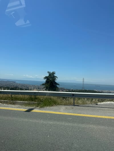 The image shows a view from a roadside in the Agios Pavlos Municipal Unit, Greece. The road, marked with a yellow line, has a metal guardrail. Beyond the guardrail is a grassy area, a tree, and a panoramic view of the city, the sea, and distant mountains under a clear blue sky. An electrical pylon is visible in the distance, over the water. Several ships are visible as well. A reflection is seen in the lower left.