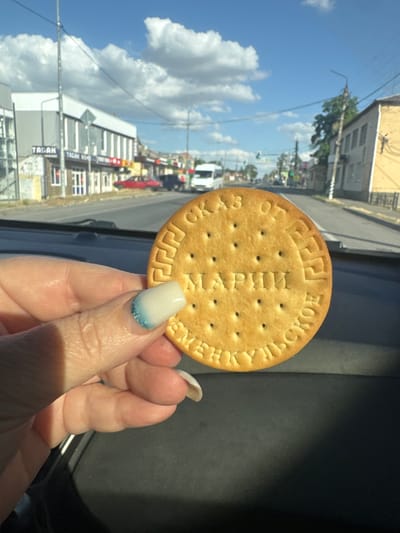 A medium shot captures a person's hand holding a round biscuit with embossed text, positioned in the foreground against a blurred background of a street scene. The biscuit, golden brown and slightly textured with small perforations, displays Cyrillic lettering. The text appears to read "МАРИИ" in the center, and "ОТЕЧЕСТВЕННОЕ" and "ВЕЧНОЕ" around the perimeter. The person's hand is fair-skinned with a manicured nail that features a white base coat with a blue glitter gradient near the tip. The background reveals a street lined with buildings, some with storefronts, and a few vehicles, including a white van and a red car. A traffic light is visible in the distance, suggesting an urban or town setting. The sky is blue with scattered white clouds. The provided location information specifies Kagal'nitskaya, Russia.
