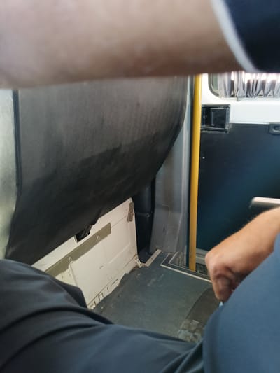 The image shows the interior of a public transport vehicle, possibly a bus, with a focus on the floor and lower interior walls. A person's legs, clad in dark blue trousers, are visible in the foreground, partially obscuring the view. To the right, another person's arm and hand are visible, suggesting they are seated or standing nearby. A yellow vertical pole, a common feature in buses for passengers to hold onto, is present. The floor appears to be dark and slightly worn. The environment is utilitarian, with visible metal panels and a black padded section of what might be a seat or wall covering. There are no clear indicators of the city of Досан, Kazakhstan, within the image itself; the context of the location would require external information. The scene is captured from a low-angle perspective, looking forward and slightly upwards.