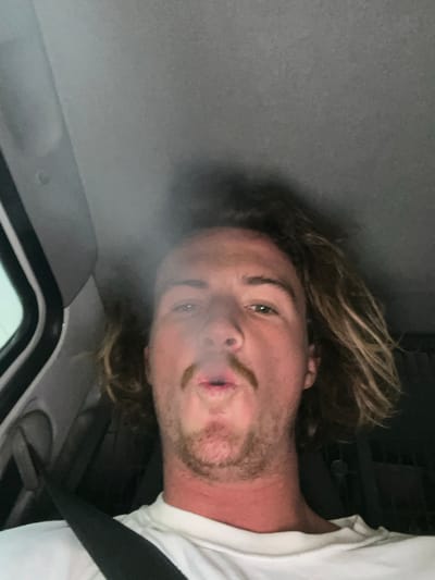 A selfie taken from a low angle captures a person inside a vehicle. The individual has light skin, wavy blonde hair, and a sparse beard with a mustache. They are wearing a white t-shirt and have a seatbelt across their chest. Smoke partially obscures their face. The interior of the vehicle is visible, with the ceiling and window frame appearing in the shot. The overall image is slightly blurry.