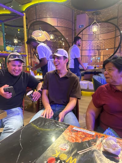 The image shows a group of people sitting at a table in what appears to be a restaurant or entertainment venue. The setting features modern decor with circular patterns and ambient lighting. The people are casually dressed and seem to be enjoying themselves.
In the foreground, two men are visible. One is wearing a black long-sleeved shirt, a baseball cap, and sunglasses pushed up on his head. He is smiling and holding a smartphone. Next to him, another man in a brown t-shirt and a white baseball cap is also smiling, looking towards the camera. Behind them, a woman in a red shirt and an older person, partially visible, are also seated at the table. Another person in a light blue t-shirt is seated further back.
The table surface is black and reflective, with a menu or placemat featuring an image of food and some text. The visible text on the placemat includes "K", "PEPSO", and phrases in Spanish that seem to relate to respecting food and a charge for wasted plates. The presence of a playful, brightly colored play area in the background, with a net structure, suggests the location might be a family-friendly venue or a restaurant with an integrated play area. The lighting suggests it could be evening or indoors with artificial lighting. The overall mood appears relaxed and convivial.