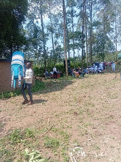 The image depicts an outdoor scene in Butali Chegulo ward, Kenya, under a clear sky. In the foreground, an adult male, dressed in a light-colored, possibly striped long-sleeved shirt, dark trousers, and brown shoes, walks from left to right, carrying a stack of approximately six to seven light blue and white plastic chairs. The ground beneath him is dry earth with sparse green vegetation, scattered leaves, and small branches. To the far left, a portion of a mud-walled structure is visible.

In the mid-distance, a gathering of approximately 20-30 individuals is observed. Most are seated on similar plastic chairs arranged in rows. A large sound system, consisting of two stacked dark speakers, is positioned near the seated group. Many individuals in the gathering are wearing light-colored clothing, with several dressed in white attire. To the far right, a light green tent or canopy is partially visible. The background is dominated by numerous tall, slender trees, forming a dense green canopy against the sky. The overall scene suggests a community event or gathering in progress.