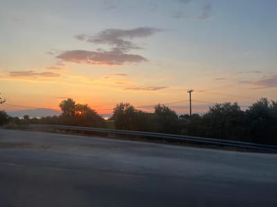 A scenic landscape captures a serene sunset over Skala Rachoniou, Greece. The sky is painted with hues of orange, pink, and blue, with scattered clouds adding texture. The sun, partially obscured by trees, casts a warm glow on the horizon, creating a tranquil and beautiful scene.

In the foreground, a paved road curves gently to the left, with a metal guardrail lining its edge. The road appears to be a quiet, rural route. Beyond the guardrail, a line of lush green trees forms a dense natural barrier, with some taller trees reaching towards the sky. The setting sun is visible peeking through the foliage of these trees on the right side of the image.

In the distance, a body of water, likely the sea, is visible, with faint outlines of mountains or landmasses on the other side. A solitary utility pole with wires is a subtle human-made element in the natural scenery. The overall atmosphere is peaceful and idyllic, suggesting a moment of calm at the end of the day. The time of day is clearly sunset, indicated by the warm, diffused light and the colors in the sky. The weather appears clear, allowing for a vibrant display of the sunset. No text is visible in the image.