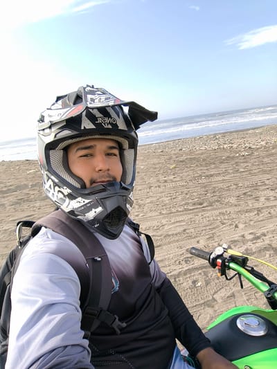A medium, eye-level shot captures a person on a green and black motorcycle on a beach, the coastline visible in the background. The individual, wearing a black and white dirt bike helmet with the brand "ONEAL" prominently displayed, a grey and black long-sleeved shirt, and a black backpack, is positioned in the foreground. The beach is sandy with visible tire tracks, and a rocky shoreline is evident further away. The sky is partially cloudy. The photo appears to be taken in Huanchaco, Peru.