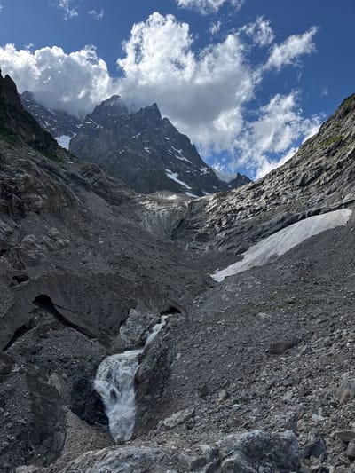 The image depicts a dramatic, high-altitude mountain landscape. In the foreground, a powerful waterfall cascades down a steep, rocky slope covered in scree and debris. The water is white and frothy, indicating a rapid descent. Above the waterfall, the terrain is characterized by rugged, dark gray rock formations interspersed with patches of snow and ice. A glacier is visible further up the valley, with its textured surface reflecting sunlight. The surrounding mountains are jagged and imposing, with sharp peaks piercing a bright blue sky dotted with fluffy white clouds. Some of these peaks are capped with snow. The overall environment is remote and wild, suggesting a glacial valley in a mountainous region. The image does not contain any people, structures, or any other elements that would identify a specific location within the city of Murshkeli, Georgia.