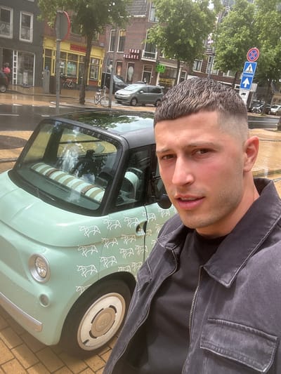 The image captures a street scene in Groningen, Netherlands, with a man in the foreground posing for a selfie. He is standing next to a light green Fiat 500, which is decorated with a pattern of white horse silhouettes. The man is wearing a dark denim jacket over a black t-shirt. In the background, several buildings line the street, including shops with yellow and brick facades. Trees are visible along the sidewalk, and traffic signs are posted on a pole. The street appears wet, suggesting recent rain. The overall impression is a candid street portrait with a focus on the man and his distinctive car.