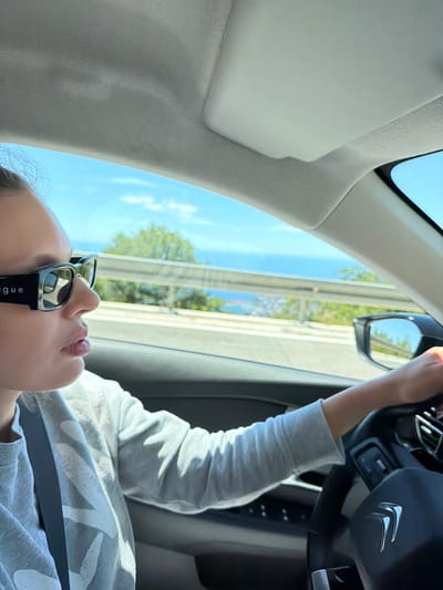 A woman wearing large, black, rectangular sunglasses with the word "vogue" visible on the side, is driving a Citroen car. She has fair skin and is looking forward. She is wearing a light grey sweatshirt with a white graphic print and a black seatbelt. The car's interior, including the steering wheel with the Citroen logo, and the dashboard are visible. Outside the car, a bright blue sky, green trees, a body of water, and a hillside town are visible, suggesting a coastal location. The image was taken from inside the car, looking forward and to the right. The location is identified as being in Stanišići, Montenegro.