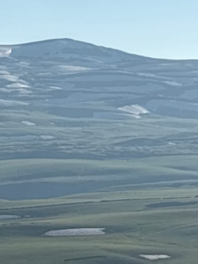 The image shows a panoramic view of rolling hills and mountains under a clear blue sky. The landscape is predominantly green with patches of snow or light-colored soil scattered across the hills. The terrain appears to be a natural, undeveloped area, likely in a rural setting. The distant mountains are hazy and appear to have snow on their peaks. The lighting suggests it is daytime, possibly morning or afternoon. Without specific landmarks or identifiable structures, pinpointing a precise location within Jermuk, Armenia, is not possible based solely on this image. The scene is serene and expansive, emphasizing the natural beauty of the topography.