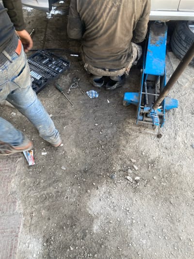 The scene depicts an active vehicle repair setting in Harasta, Syria. Two individuals are present, engaging in mechanical work on a light-colored vehicle, a portion of which is visible at the top of the frame. One individual is observed from behind, kneeling or crouching beneath the vehicle, wearing a dark, possibly brown, long-sleeved shirt and dark trousers, with dark footwear. Their hands are out of view, suggesting active work on the vehicle's underside. A second individual is partially visible on the left, from the waist down, wearing light blue jeans, a brown belt, and dark athletic shoes, standing near the work area.

The vehicle is supported by a prominent blue hydraulic floor jack, positioned beneath its right side, with a tire visible adjacent to the jack on the far right. An open black tool kit, filled with various sockets and wrenches, is situated on the ground near the standing individual. Other tools scattered on the dirt include a green-handled screwdriver and a pair of silver-colored pliers. A crumpled blue and white checkered cloth or rag lies near the kneeling individual. The ground consists of loose dirt, gravel, and patches of worn reddish-brown pavement, exhibiting significant soiling consistent with grease and oil from vehicle maintenance. A discarded food wrapper is visible near the standing individual's feet.