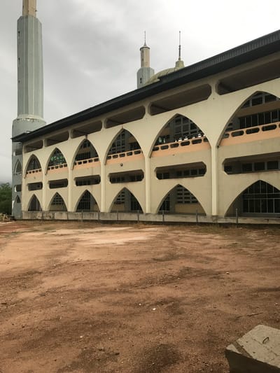 The image shows a section of the Obafemi Awolowo University Mosque in Ile-Ife, Nigeria. The architectural style is a series of repeating arches, with windows in alternating arch segments. The building is beige with pale peach accents. A minaret extends vertically from the structure. The foreground is composed of rough, reddish-brown dirt and debris. The sky is overcast.