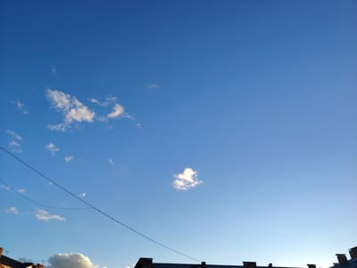 The image shows a clear blue sky with scattered white clouds. In the foreground, the rooftops of several buildings are visible, silhouetted against the sky. A thin black wire, likely a power line, stretches across the frame from left to right. The location is identified as the city of Border Gyumri-Akhuryan, Armenia.