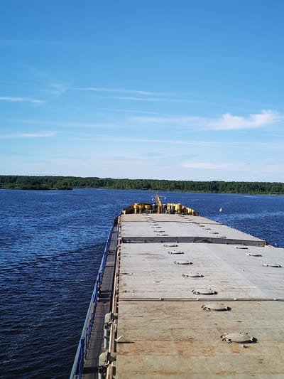 This media file depicts a bright, sunny day on a wide, calm river, likely in Даргун, Russia. The perspective is from the deck of a large, flat-bottomed barge, looking forward and slightly to the left. The barge's deck is weathered and industrial, covered with numerous circular hatches, some of which are raised. A blue railing runs along the left side of the barge, separating the deck from the deep blue water.
The river stretches out before the viewer, meeting a dense, green treeline on the opposite bank. The trees form a lush and unbroken horizon under a clear, pale blue sky dotted with thin, wispy clouds. The water exhibits a gentle ripple, suggesting a slow movement of the barge. In the distance, a white buoy is visible on the water.
Towards the front of the barge, a section with yellow structures is present, possibly part of the barge's cargo or equipment. The overall atmosphere is serene and tranquil, indicative of a peaceful journey on the water under good weather conditions. The time of day appears to be mid-morning or early afternoon, given the bright sunlight and clear sky. No people are visible in the frame, and there is no visible text.