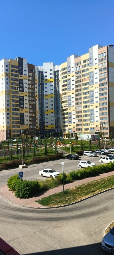 The image depicts a residential area in Zonalnaya Stantsiya, Russia, under a clear blue sky. In the foreground, a curved asphalt road leads into a parking lot filled with several parked cars, including white sedans, a white SUV, and a silver sedan. The parking lot is bordered by a paved walkway and lush green bushes. Behind the parking lot, a playground with swings and slides is visible amidst manicured green spaces and young trees. The background is dominated by a large, multi-story apartment building complex. The building features a modern design with a segmented facade, alternating between yellow, white, grey, and brown panels. Numerous windows are visible across the building's exterior, with some appearing to have curtains drawn. The scene suggests a typical day in a suburban residential environment.