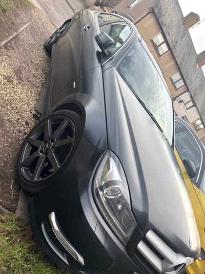 A matte dark grey Mercedes-Benz C-Class sedan is parked at an angle on a residential street verge in Montrose, United Kingdom. The vehicle features dark grey multi-spoke aftermarket alloy wheels with Mercedes-Benz center caps, and red brake calipers are visible behind the front wheel. Its front end displays clear lens headlights, a prominent grille with a large Mercedes-Benz emblem, and lower bumper sections with chrome accents around what appear to be fog light housings. The car is positioned with its passenger side partially on a patch of grass and a light-colored gravel strip. A paved surface, possibly a sidewalk or road edge, runs alongside the gravel. Behind the Mercedes, the front portion of a yellow compact car is partially visible. In the background, typical two-story residential buildings with brown/tan facades, multiple windows, and dark-tiled or slated pitched roofs line the street. The sky is overcast. No individuals or specific actions are visible. The scene depicts a parked vehicle in an urban residential setting.