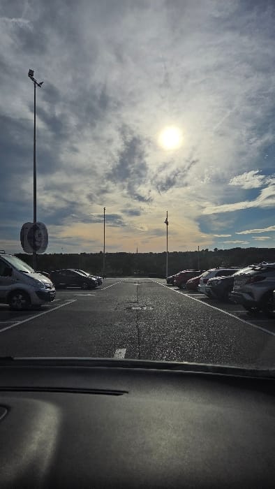 The image presents an outdoor asphalt parking lot in Paterna, Spain, observed from the interior of a vehicle, looking over a dark dashboard. The sky is partially cloudy with a prominent, bright sun positioned in the upper right-center. White and grey clouds dominate the sky, with faint blue and orange hues near the horizon line. Contrails are visible in the upper atmosphere.

A tall, dark utility pole is situated on the left side of the frame, featuring a street light fixture and a circular, partially obscured sign that appears to be a "P" or an abstract symbol. Another slender pole stands further down the center of the lot.

On the left, a silver van is parked, with additional dark-colored vehicles parked further down the row. On the right, a row of multiple parked vehicles, including several SUVs and sedans of varying colors (red, white, dark grey, silver), extends into the background. The asphalt pavement displays white painted lines marking parking spaces and lanes.

The background consists of a low, dark green landscape, likely a treeline or hills, stretching across the horizon. No individuals are present. The lighting conditions suggest either early morning or late afternoon.