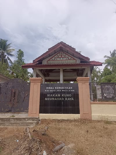 A weathered monument stands at the entrance to a site identified as "MAKAM KUNO MEUNASAH RAYA" (Ancient Cemetery of Meunasah Raya) in Meunasah Raya, Indonesia. The monument is a dark tile plaque with white lettering, flanked by two thick, coral-colored pillars. Above the plaque, a structure with a red, multi-tiered roof, supported by white pillars, offers shade. Intricate, dark metal fencing with decorative patterns surrounds the area. Tropical trees, including palm trees, are visible in the background under a cloudy sky. The ground in front of the monument is covered in dry, brown grass and scattered debris, including branches and roots. The scene is static, with no people or animals present.