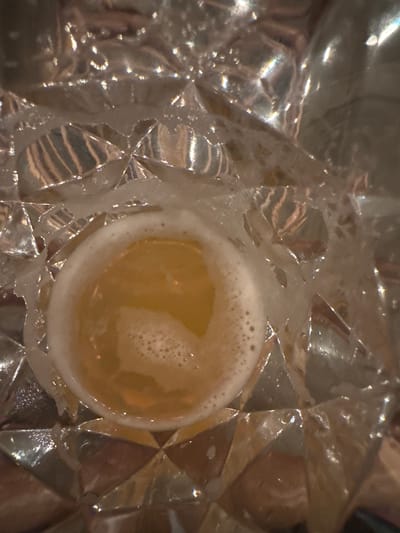 A close-up, overhead shot shows a glass of amber-colored liquid with a foamy head, resting on a textured, reflective surface. The surface appears to be made of crystal or glass with a diamond-like pattern. The liquid is a warm, golden hue and the foam is white with small bubbles. The lighting creates highlights and shadows across the scene, emphasizing the reflective qualities of the glass and the texture of the surface. No people or specific environmental details of Chuguyevka, Russia are visible in this image.
