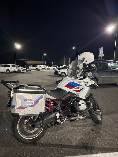 A white BMW R1250GS Rallye motorcycle is parked in an outdoor parking lot at night. The motorcycle has a white helmet resting on its handlebars. The background shows several parked cars and streetlights. The location is identified as the city of Privolzhsky, Russia. The ground is asphalt, and there are painted white lines marking parking spaces. The sky is dark.