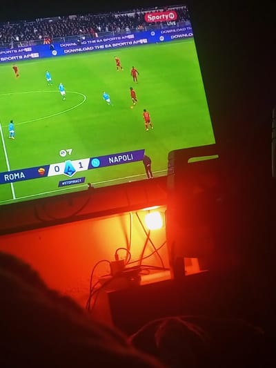 A television screen displays a soccer game between Roma and Napoli, with Napoli leading 1-0. The game is branded with "Sporty TV LIVE" and features an advertisement to "Download the EA Sports App."  A hashtag "#STOPIRACY" is shown below the score. The room is dimly lit, illuminated by a warm red light that highlights cables, a charging block, and a partially visible object resembling a speaker. The bottom of the frame captures a blurry glimpse of a person.