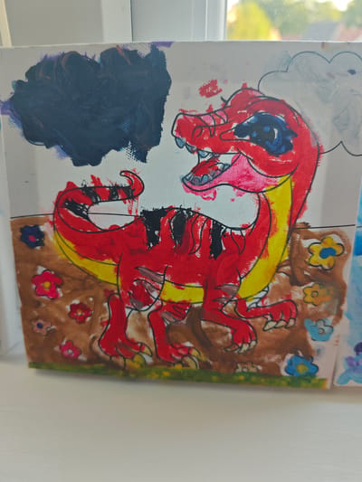 A painting on canvas depicts a red cartoon dinosaur with yellow accents on its neck and tail. The dinosaur is standing on brown ground dotted with colorful flowers. Above the dinosaur are dark blue and grey clouds. The painting is on a windowsill, with a blurred background of trees and buildings visible through the window. The location is Diss, United Kingdom.