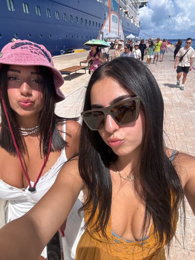 The image displays a close-up selfie of two young adult females on a sunny pier in San Miguel de Cozumel, Mexico.
The woman positioned on the left wears a pink bucket hat with the text "CAYMAN ISLANDS B.W.I." visible, a white sleeveless top with lace trim, and multiple white bead necklaces. A red cord hangs around her neck. Her dark hair frames her face, and her lips are pursed.
The woman on the right wears large, dark-framed rectangular sunglasses, a gold-colored chain necklace with a small pendant, and an ochre-colored sleeveless top with lace detail over a light blue garment. Her dark hair falls around her shoulders, and her lips are also pursed. Both subjects are looking towards the camera.
In the background, a large blue multi-deck cruise ship is docked at the pier. The pier itself is paved with light-colored material, and several wooden benches are visible. Numerous individuals, identified as pedestrians or tourists, are walking on the pier. One individual in the mid-ground uses a multi-colored umbrella for sun protection. The sky is blue with scattered white clouds.