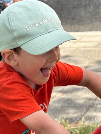 A close-up, profile shot captures a young boy wearing a light green baseball cap with "LET'S HAVE FUN" embroidered in light pink lettering. He is also wearing an orange t-shirt. The boy is looking to his right with his mouth open and tongue sticking out, as if mid-laugh or speaking. His right ear is visible above the cap, and his skin appears flushed, suggesting activity or warmth. The background is out of focus, showing a textured, greyish-brown surface, likely pavement or concrete, with hints of green grass at the bottom right. The scene is outdoors, possibly in a park or playground area in the city of Vinarsko, Bulgaria.