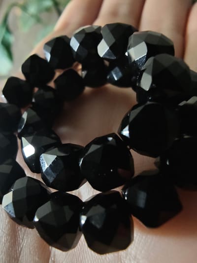 A close-up, slightly angled shot shows a hand holding a black beaded bracelet. The beads are faceted and appear to be made of a dark, reflective material like onyx or obsidian. The hand is positioned to cradle the bracelet, with fingers slightly curled. The background is softly blurred, suggesting an indoor setting with diffused lighting. No specific location in Kursk, Russia, is identifiable from the visual information provided.