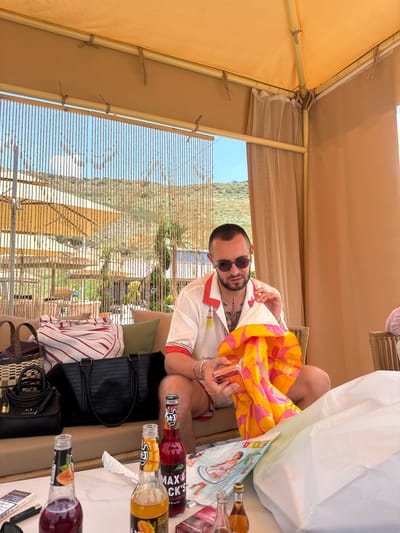 In an outdoor setting, a man with sunglasses and a beard, wearing a white collared shirt with red trim, sits on a couch. He is holding an orange and pink patterned bag and a phone. Nearby, there are two bottles of what appears to be alcohol, one labeled "MAX & CKS Y & ALMOND 7% ALC/VOL". A white bag with images of people on it is also present, along with several other bags, including a black crocodile-textured tote bag and a smaller black bag with a gold clasp. The scene appears to be under a tan canopy, suggesting a shaded outdoor area. In the background, there is a view of hills and greenery, indicating a sunny day, possibly in the afternoon, given the shadows. The overall mood seems relaxed and leisurely. Visible text includes "AX", "EXCHANG", "CHAPMAN", "MAX & CKS", "Y & ALMOND", "FRIDS", "Greetings", "ESSE", and "INTE". The location is identified as Ptghni, Armenia.