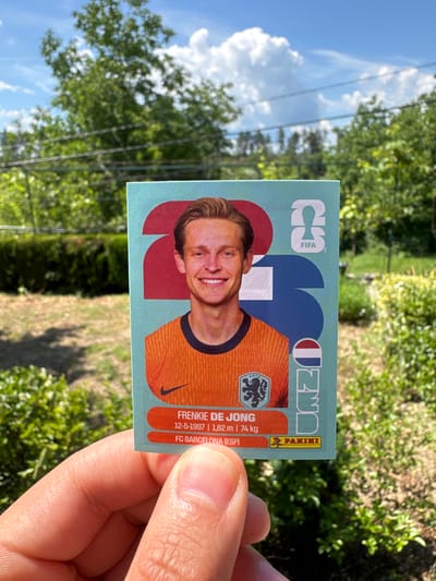 A person's hand holds a soccer sticker of Frenkie de Jong. The sticker features a portrait of the player in an orange jersey, with the Dutch flag and the number "2" and "1" visible in the background. Text below the player identifies him as "Frenkie de Jong," his birthdate "12-5-1997," height "1,82 m," weight "74 kg," and club "FC BARCELONA (ESP)." The Panini logo is also present. The sticker is held against a backdrop of green trees and a blue sky with white clouds. The image appears to be taken outdoors, possibly in a garden setting. The presence of the sticker and the natural environment suggests a casual, recreational context. There is no discernible connection to Prisovo, Bulgaria.