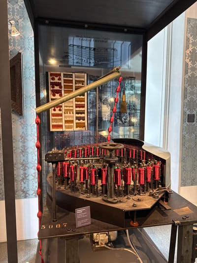 Here's a description of the image:

The image captures a vintage "Formenflecht Maschine" (shape braiding machine) at a museum display. This machine, from the Mech. Posamenten - Fabrik Gustav Gerster, likely produced decorative passementerie items. The machine features rows of bobbins, each wound with red thread, that intertwine to create intricate patterns. A sign near the machine provides information in German, indicating its function: "Mit 72 Büchsenklöppeln werden Holz und Pappformen umflachter" (With 72 bobbin clappers, wooden and cardboard forms are braided around).

The machine is behind a glass case. A metal tube structure is attached to it. In the background of the display is an illustration and examples of the kinds of products it would have made. The display is next to a patterned blue and white wallpaper. The number "5007" is painted on the machine.

The museum is likely located in Biberach an der Riß, Germany, given the German text on the signage and the machine's historical context within the country's textile industry.